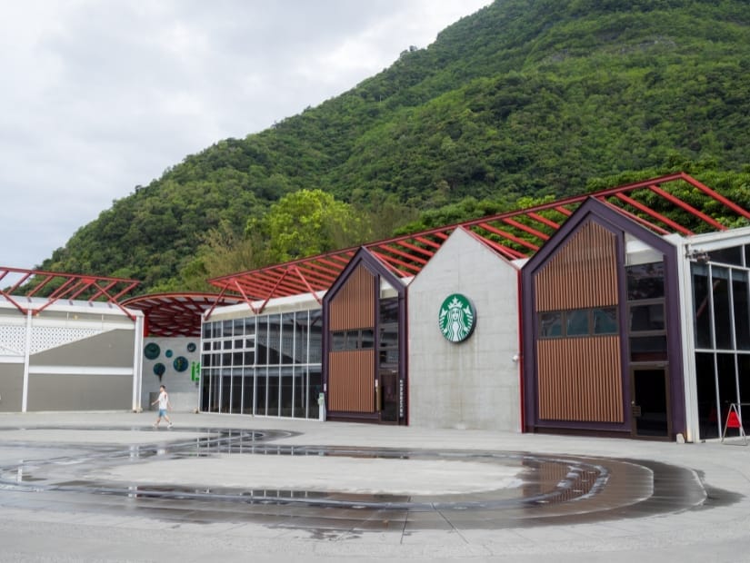 The exterior of a large cement and wood Starbucks in Hualien with water fountains in a square in front of it and green mountains behind it