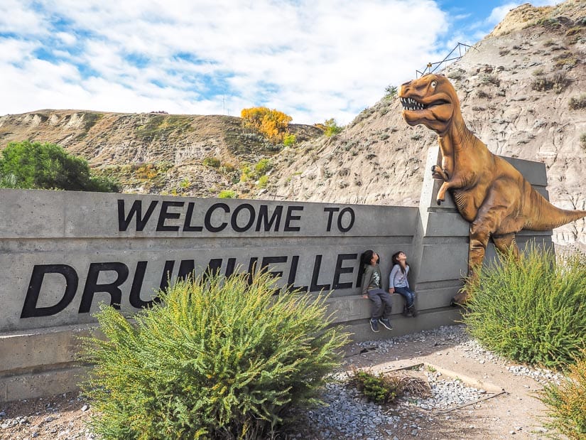 Two kids standing below a dinosaur on a "Welcome to Drumheller" sign