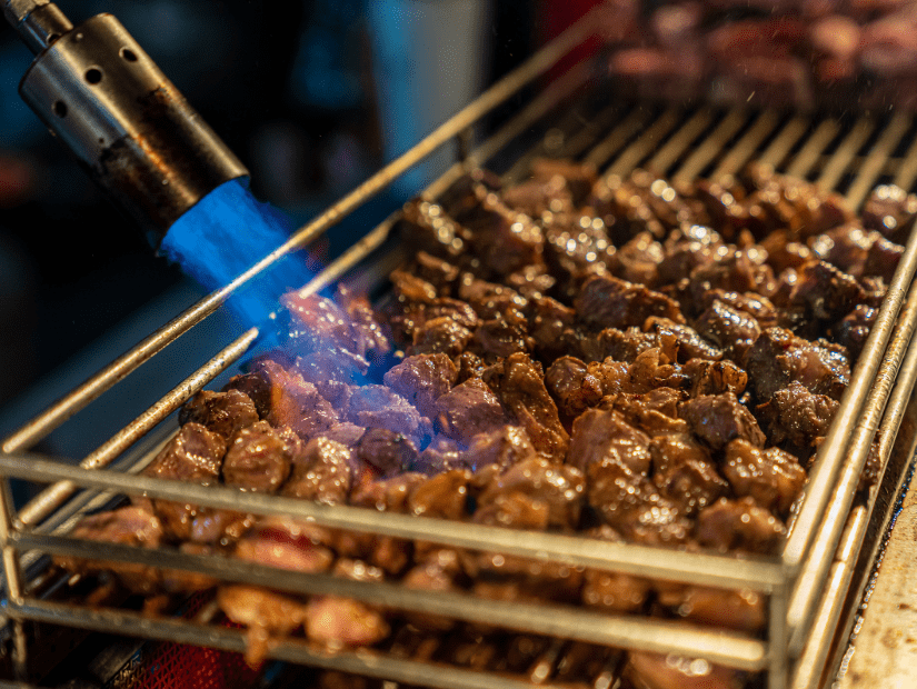 A blow torch shooting flames at some cubes of beef on a grill