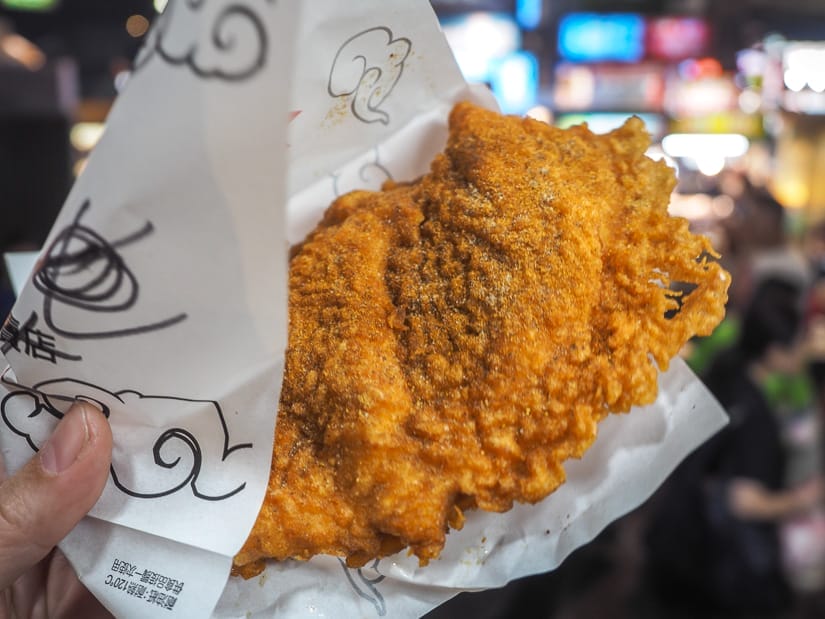 A hand holding up a huge battered and deep fried chicken fillet in a paper wrapper in a night market