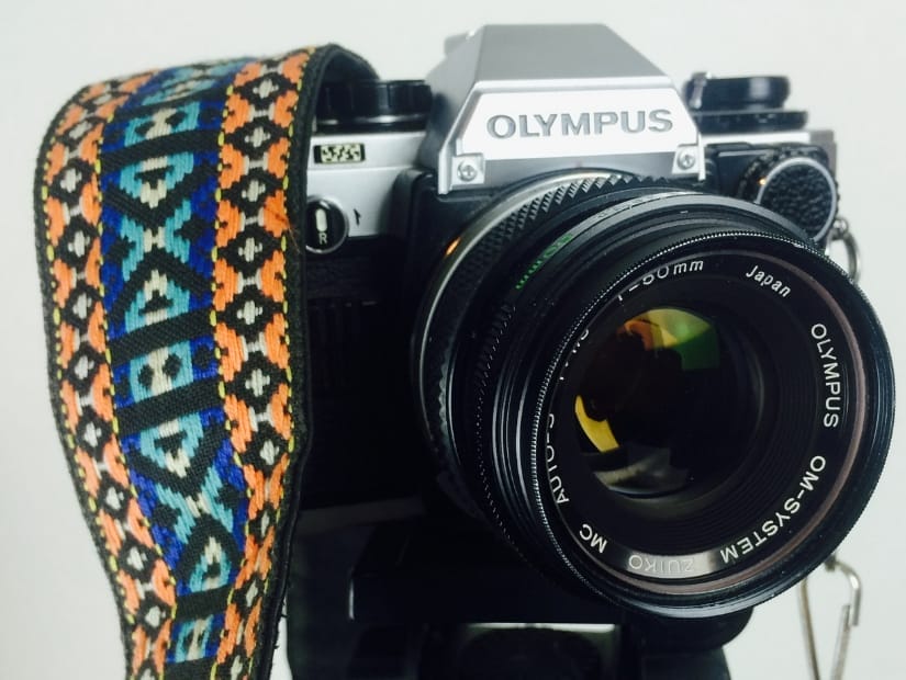 The front of an old-fashioned looking Olympus camera with a colorful camera strap wrapped around the side of it.