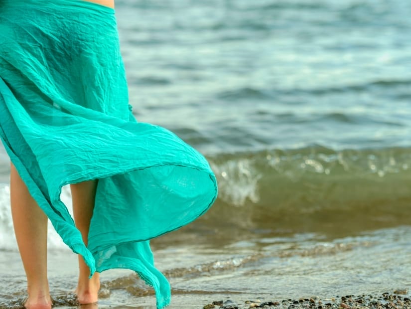 A person's lower half viewed from behind, wearing a green sarong which is blowing in the wind, while standing on a shore facing the waves