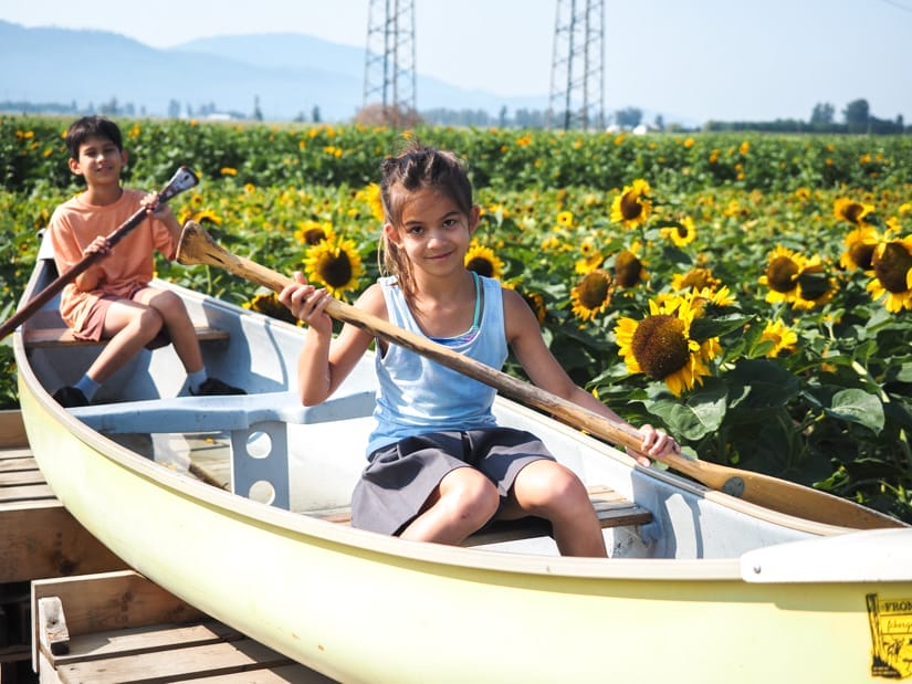 Two kids pretending to paddle in a canoe that's on land with a sea of sunflowers beside