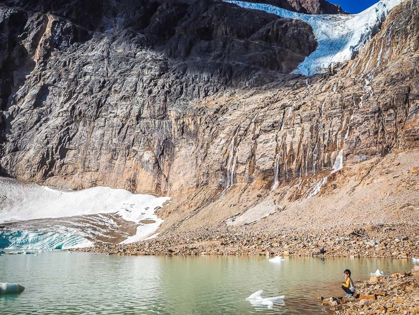 A kid kneeling by the shore of Cavell Pond with a glacier on the mountain above him