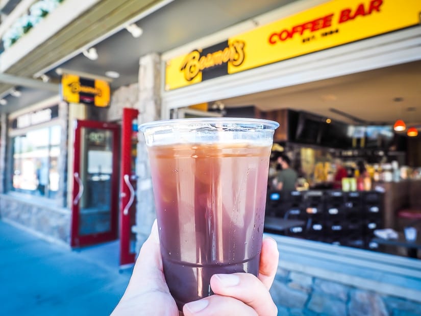 A hand holding up an iced american in front of Beamers, the best cafe in canmore