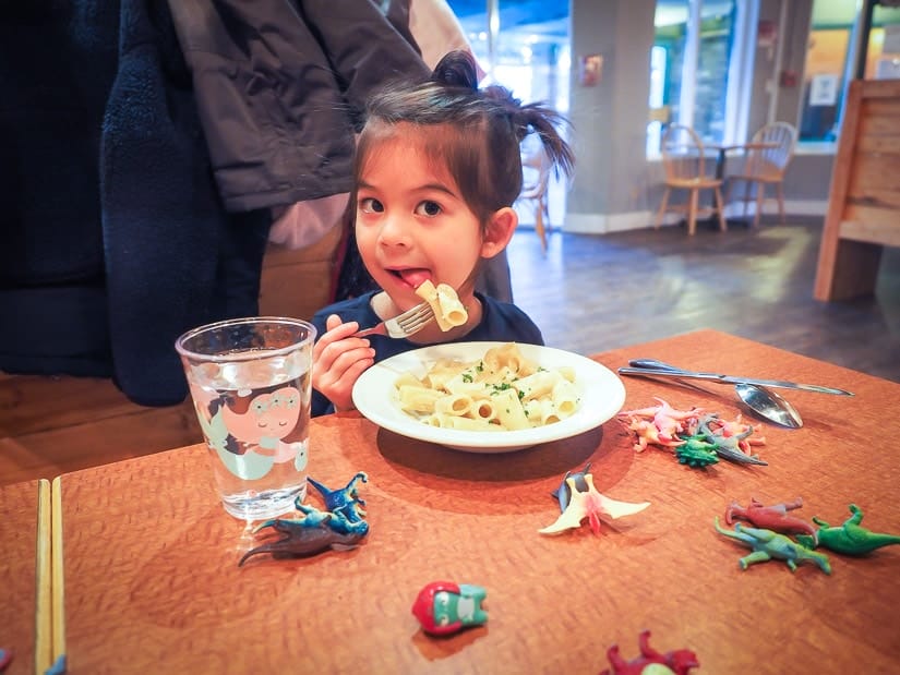 A girl eating pasta with some toy dinosaurs on the table