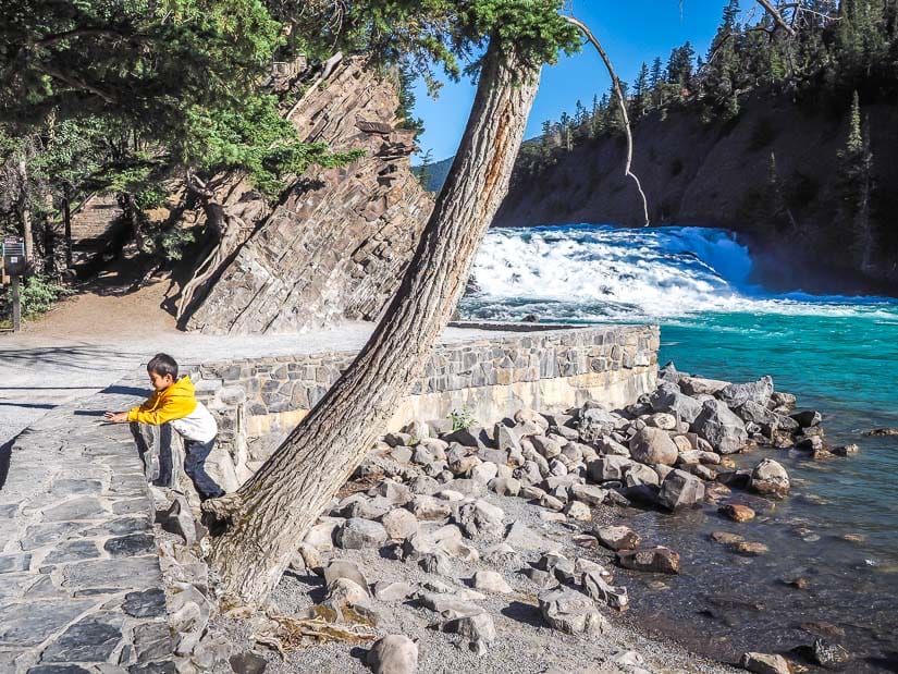 A kid climbing a stone wall with Bow Falls in the background