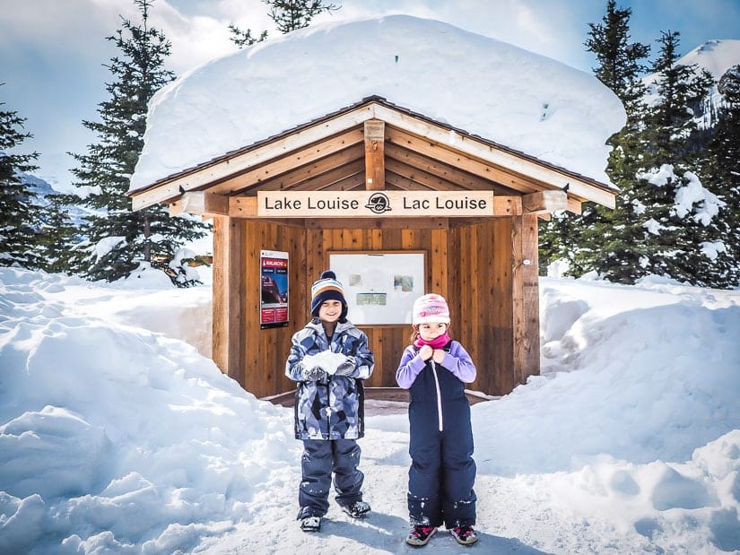 Two kids in a snowy scene in front of a sign that says Lake Louise