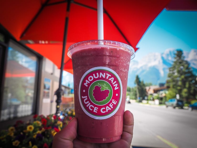 A hand holding up a red smoothie in front of Mountain Juice Cafe in Canmore