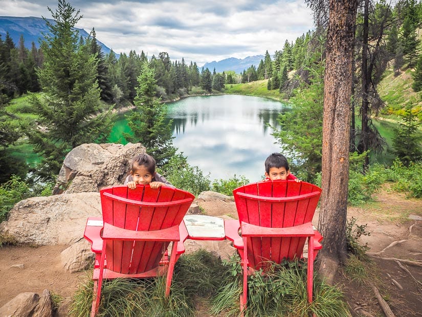 Two kids peeking over the back of two large red chairs, with a beautiful lake in the background.