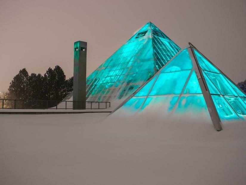 Two glass pyramids of the Muttart Conservatory lit up with blue lights and surrounded by snow