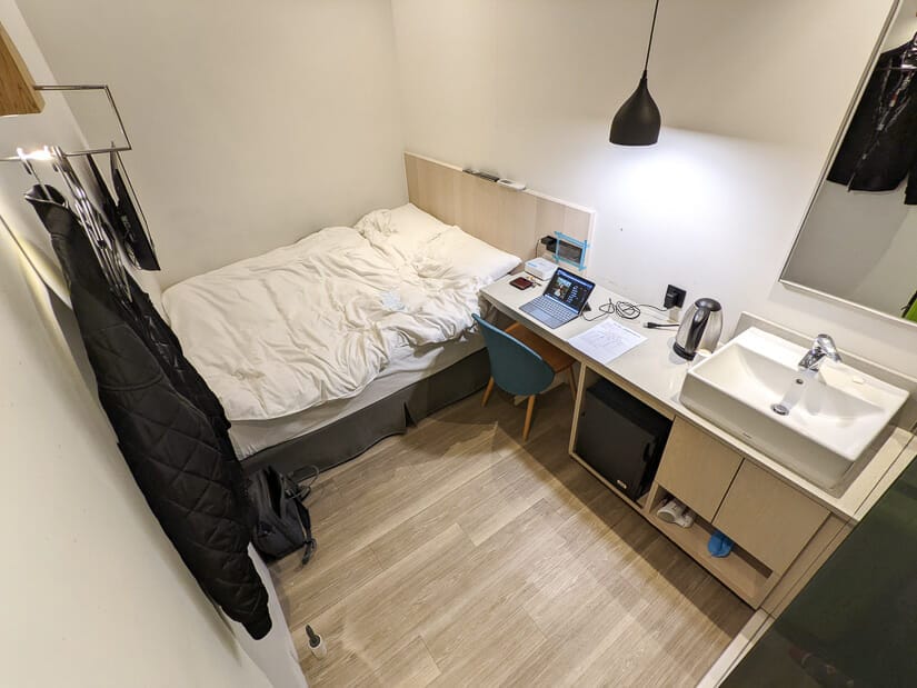 View of a very small quarantine hotel room in Taipei, including bed, desk with computer, and clothes hanging on hooks, with no window on wall.