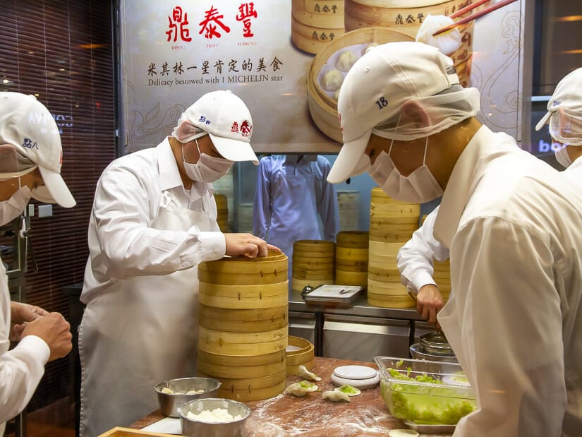 Some chefs in white clothes making xiaolongbao and putting them in baskets in a kitchen and Din Tai Fung