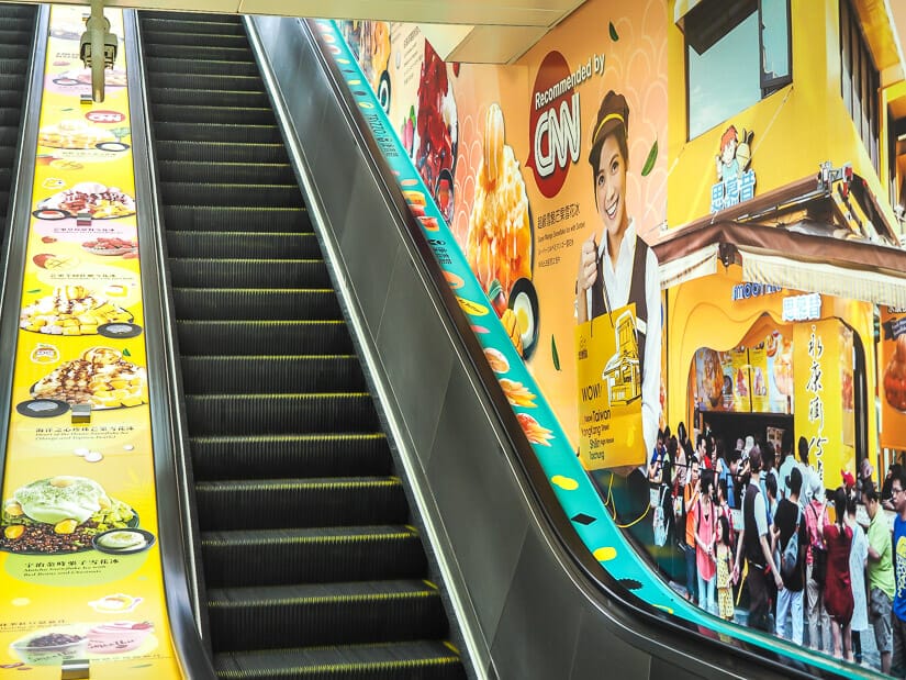 A escalator leading up out of Dongmen MRT station in Taipei, with huge orange ads for shaved ice covering the walls.
