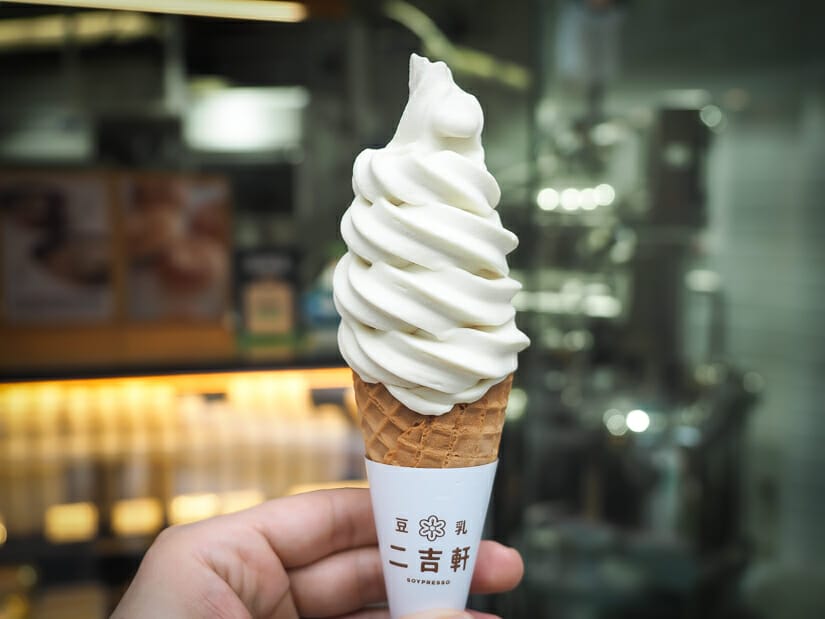 A hand holding up a white soft serve ice cream cone with Soypresso shop on Yongkang Street in the background