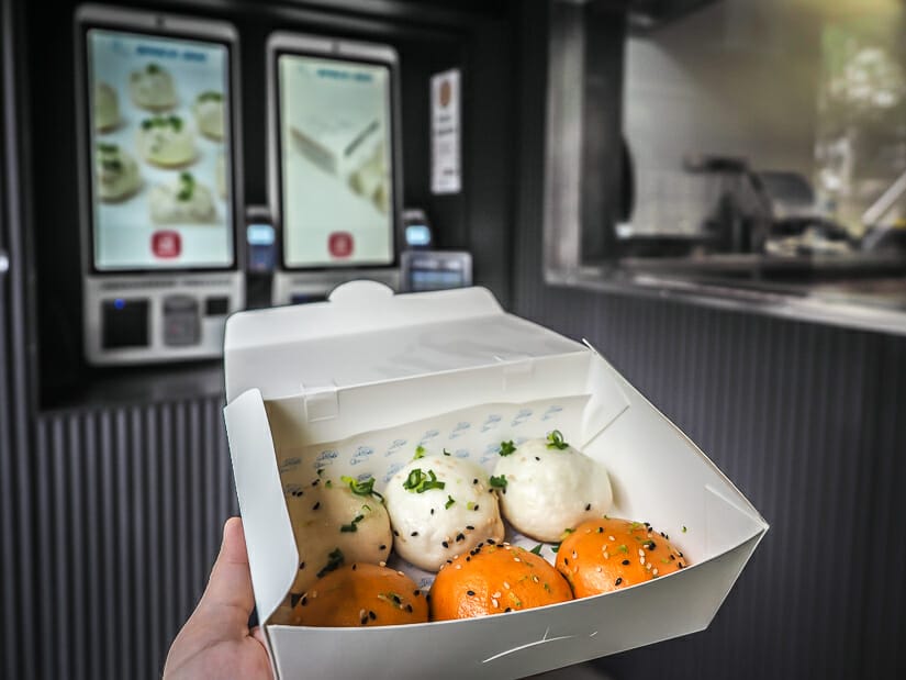 A hand holding up a take-away box with three white and three orange buns in it, with screens for ordering and a take-out window in the background