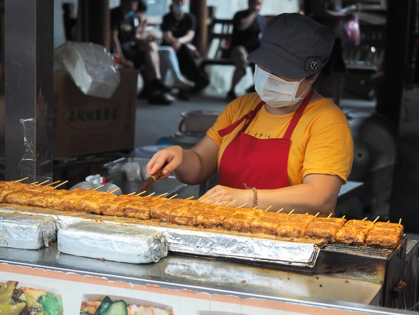 A female food vendor in a red apron preparing several orders of stinky tofu in Shenkeng
