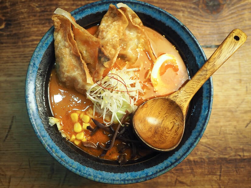 A bowl of ramen on a table with a wooden spoon and crispy wontons on top