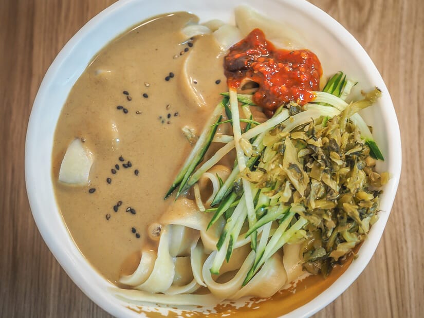 A bow of thick noodles with brown sesame sauce, cucumber slices, and spicy sauce on top