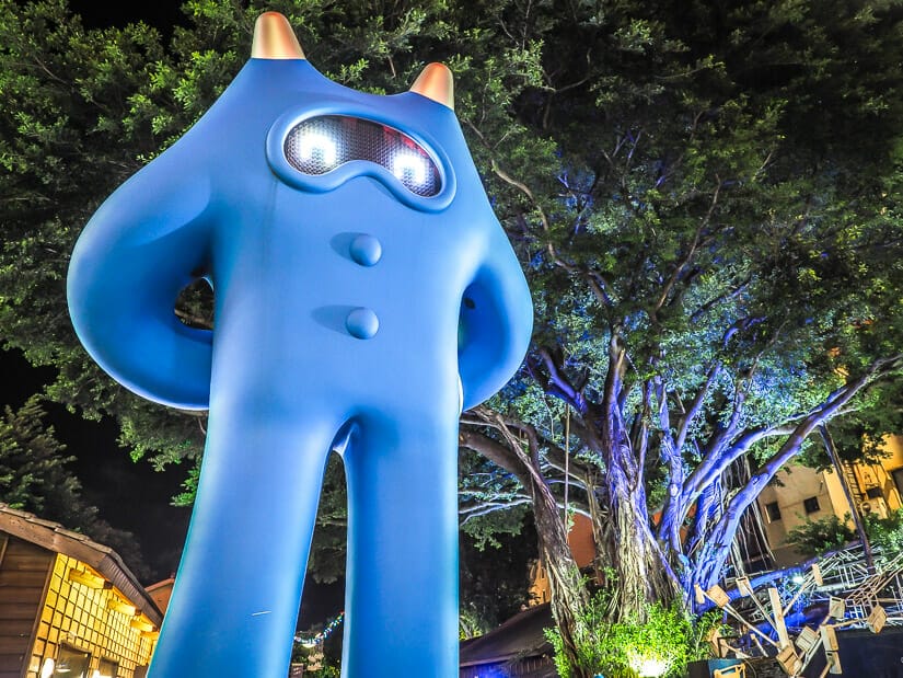 A giant blue robot standing on the street with a tree lit up with blue lights behind it