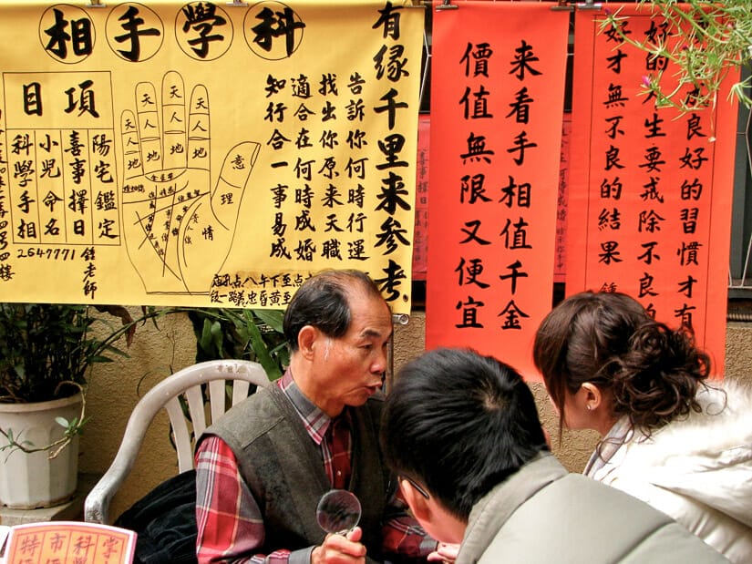 A fortune teller man sitting at a table advising two people, with red and yellow banners behind him showing Chinese characters and a hand with the different fortune lines