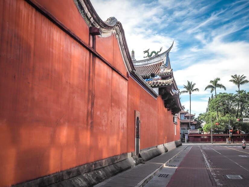 A long red wall beside a street, with one person riding a scooter on the street