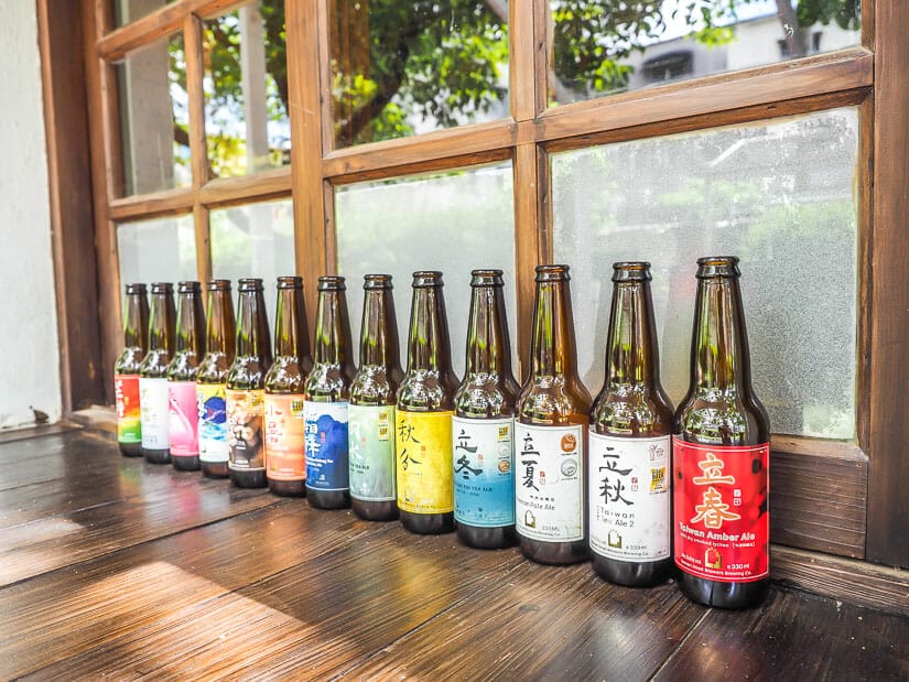 A row of beer bottles on a wooden shelf in front of a window at Anping Treehouse Cafe