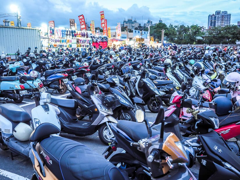 A whole bunch of parked scooters in a parking lot with the lights and stalls of Ta-Tung Night Market in the background