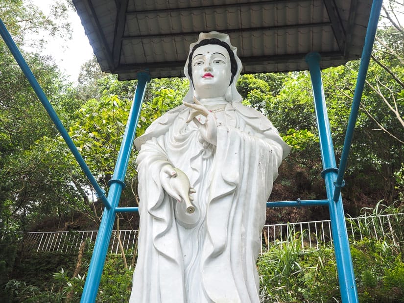 A white Guanyin goddess statue in the forest with small roof over its head