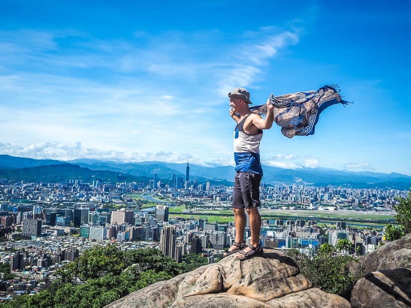 Me standing on top of a rock at the peak of Jinmianshan trail in Taipei, holding my arms out with a blue sarong blowing in the wind