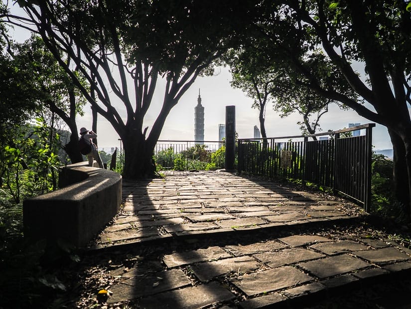 A stone lookout platform with long shadows from the trees, a person standing on the side taking a photo, and Taipei 101 visible beyond