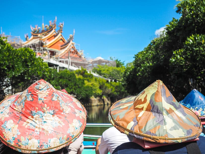 Two passengers on Sicao Green Tunnel boat tour wearing farmer's hats, with a temple visible at the top left