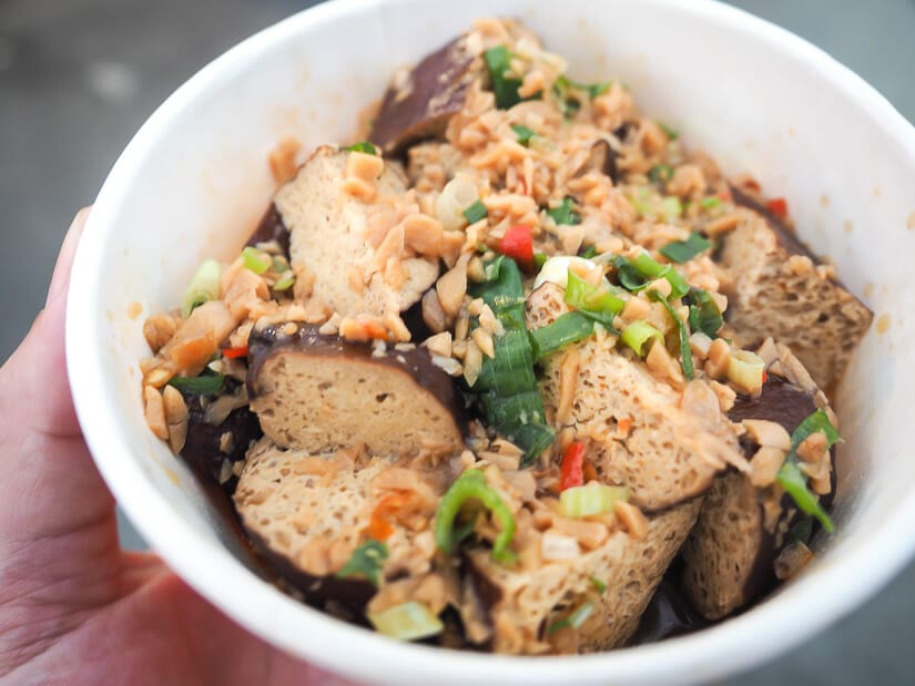 A hand holding a white paper bowl filled with tofu and toppings