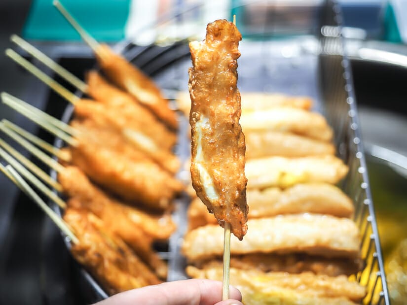 A hand holding up a stick of deep fried swordfish, with more behind it and a vat of oil