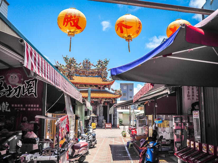 A lane with two yellow lanterns hanging above it and leading to a small colorful temple