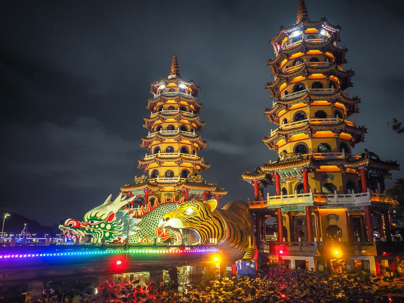 A bridge leading to a Dragon and Tiger statue with their mouths open and two tall pagodas behind them at night with lights