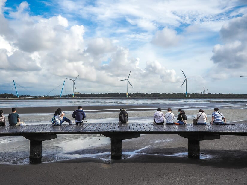 A few couples and groups of friends sitting on the dock at Gaomei Wetlands and looking at the wind turbines in the distance