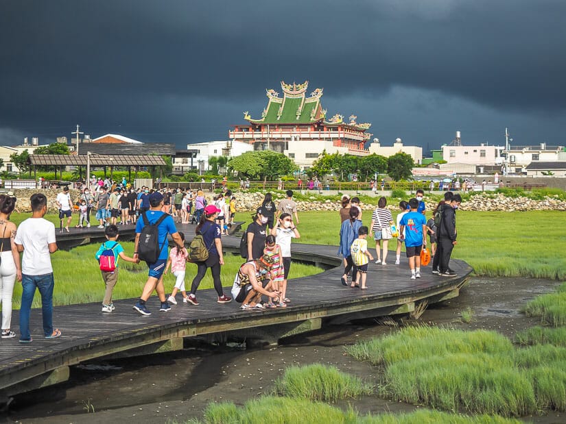 A dock with lots of people on it and a temple in the background, and the people are looking down at the wetlands of Gaomei in Taichung
