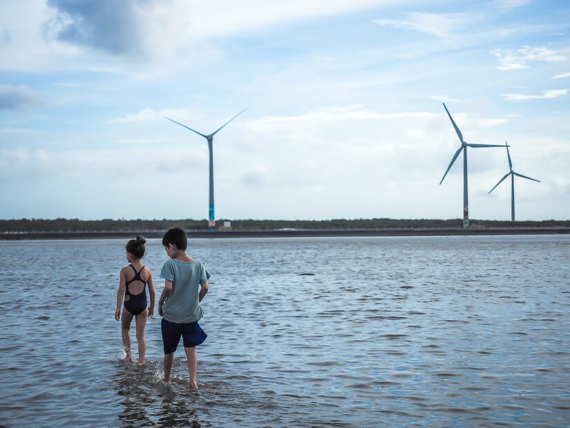 Two kids standing in shallow water at Gaomei Wetlands, with wind turbines in the distance