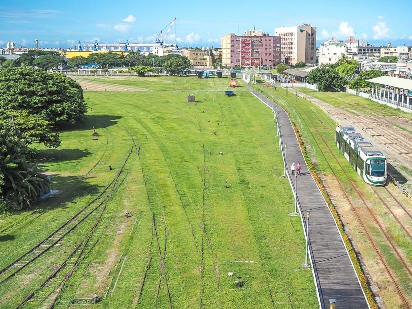A green field with old railway lines, pedestrians walking along a wooden boardwalk, and LRT driving through at Hamasan Railway Cultural Park in Kaohsiung