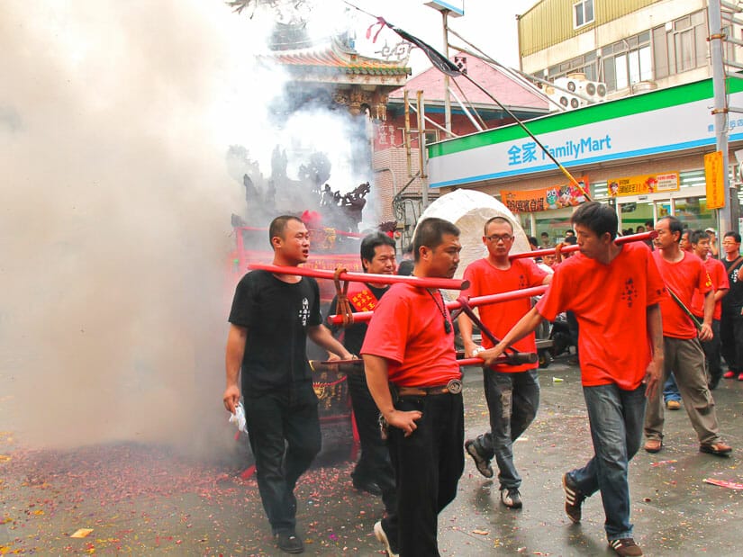 Some men in red T-shirts pulling a palanquin with lots of smoke from firecrackers around them and a Family Mart store behind them