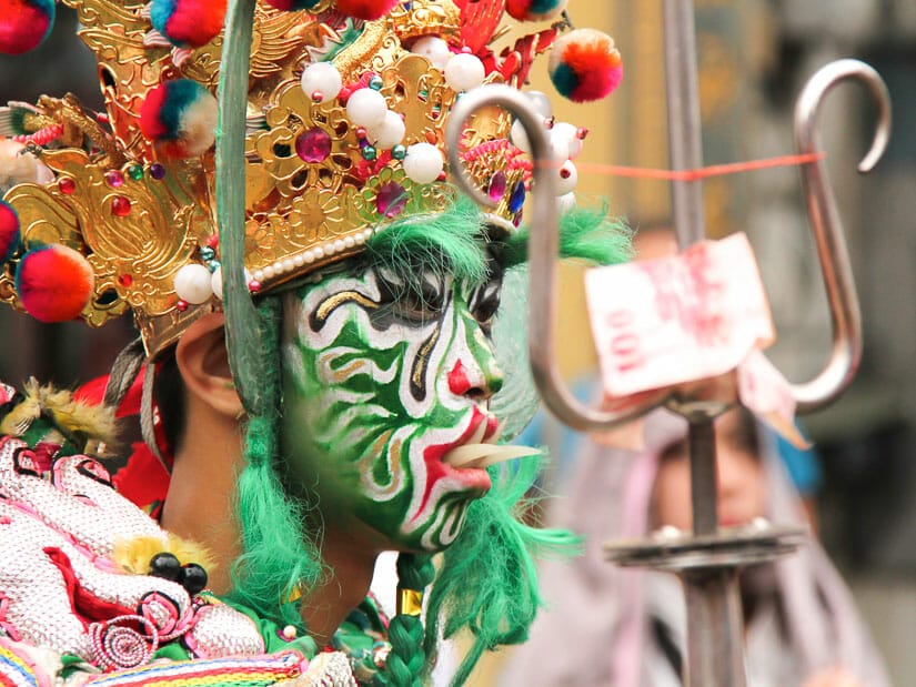A Taiwanese traditional temple performer with his face painted green, fake sharp teeth sticking out, and hold a trident with a Taiwanese 100 dollar bill on it