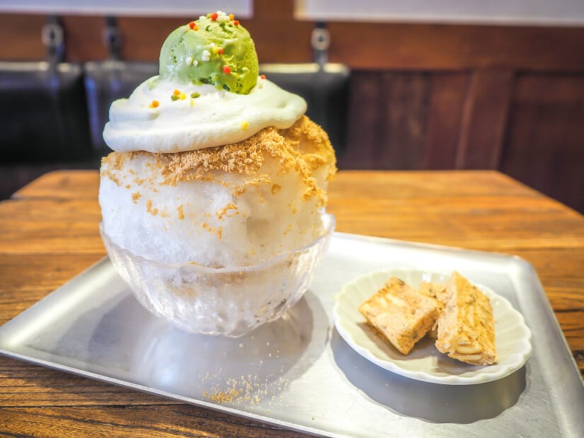 A metal tray with a shaved ice dessert and a plate of cookies on it, atop a wooden table in a cafe on Lukang Old Street