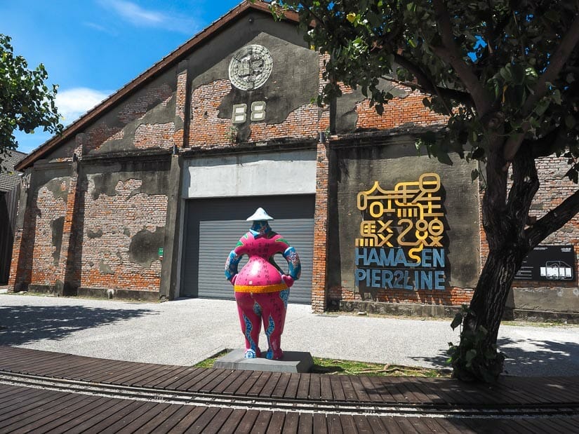 A statue standing in front of a warehouse at Penglai area of Pier 2 Arts Center and a small train track line in the foreground