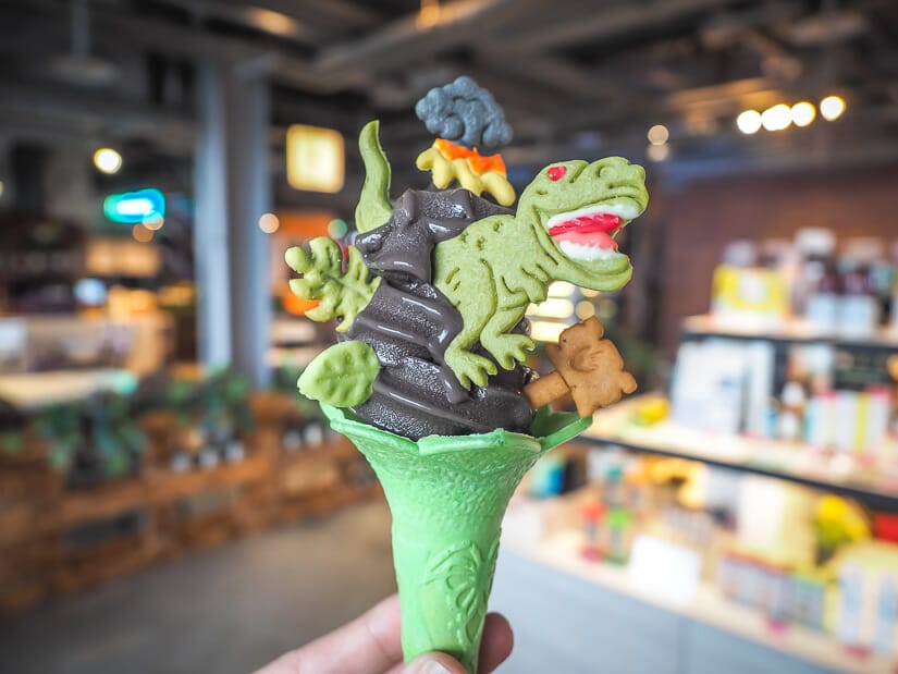 A hand holding up a green ice cream come with gray ice cream and a dinosaur cookie on it