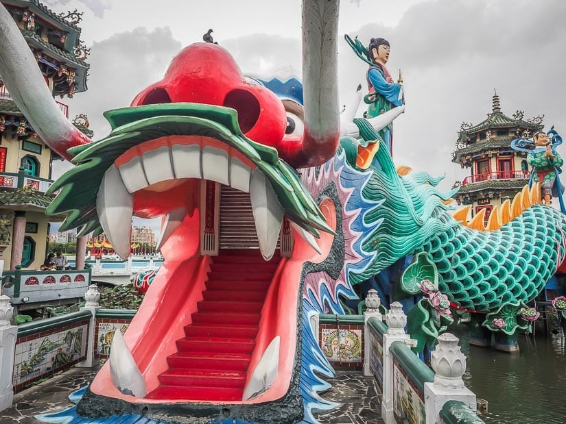 A large colorful dragon statue with its mouth open and a tunnel inside its mouth and Buddhist statues in the background