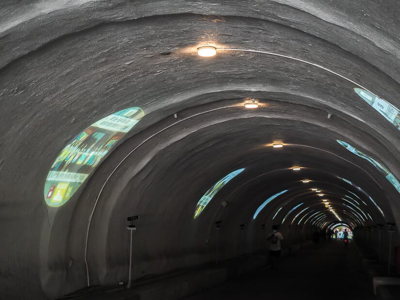 A dark, long tunnel with lights running through it and a few people walking and cycling through