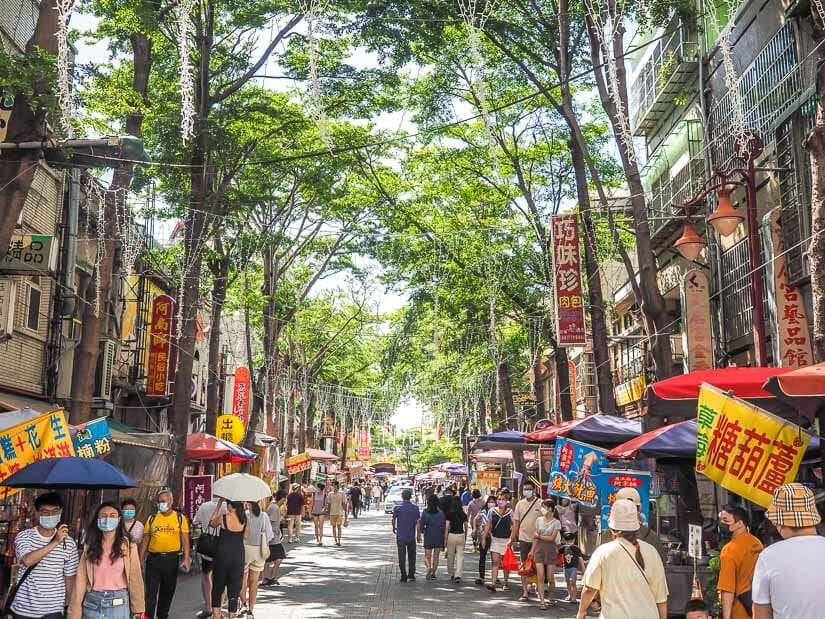 A busy street in Lukang with food stalls on either side, many pedestrians, and a canopy of trees above