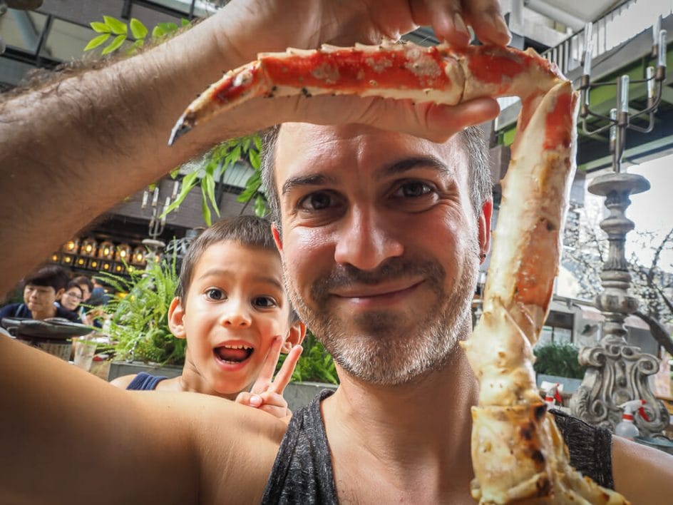 Me holding up a king crab leg at Addiction Aquatic's outdoor grilled seafood patio, with my son in the background