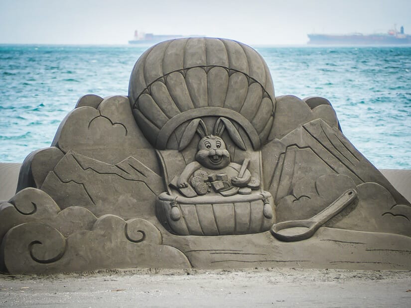 A sand sculpture of a bunny in a hot air balloon at the Cijin Beach Sand Castle Festival with the sea and ships in the background